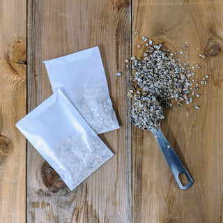 Two open white paper bags of vermiculite on a wooden surface, with some vermiculite scattered on the table beside them.